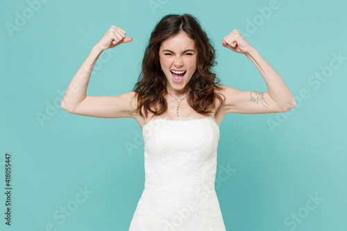 Cheerful strong bride young woman 20s in beautiful white wedding dress showing biceps muscles on hands isolated on blue turquoise color background studio portrait. Ceremony celebration party concept. © ViDi Studio