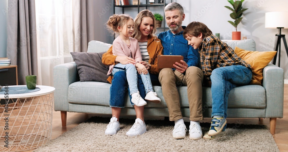 Caucasian happy cute family with children resting on sofa in cozy living room at home browsing on tablet device watching cartoons or searching internet, parents and kids spending time together