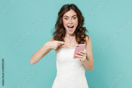 Surprised bride young woman 20s in white wedding dress pointing index finger on mobile cell phone typing sms message isolated on blue turquoise background studio. Ceremony celebration party concept.
