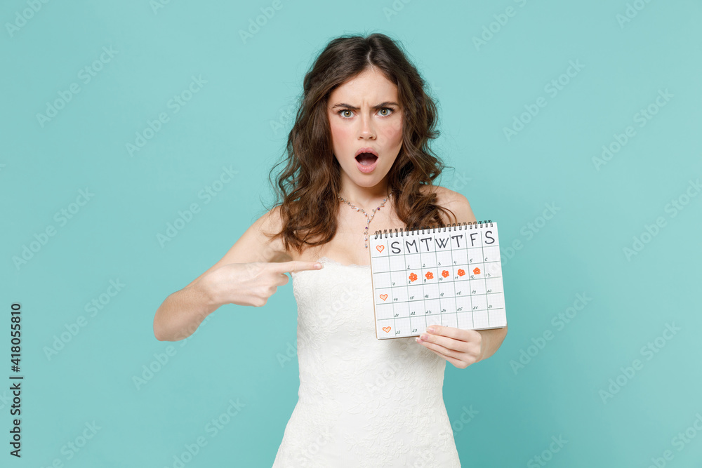 Shocked worried bride woman in white wedding dress pointing finger on female periods calendar for checking menstruation days isolated on blue turquoise background. Ceremony celebration party concept. Shocked worried bride woman in white wedding dress pointing finger on female periods calendar for checking menstruation days isolated on blue turquoise background. Ceremony celebration party concept.