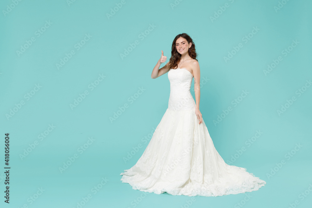Full length of smiling tender bride young woman in white wedding dress showing thumb up like gesture isolated on blue turquoise color background studio portrait. Ceremony celebration party concept. Full length of smiling tender bride young woman in white wedding dress showing thumb up like gesture isolated on blue turquoise color background studio portrait. Ceremony celebration party concept.