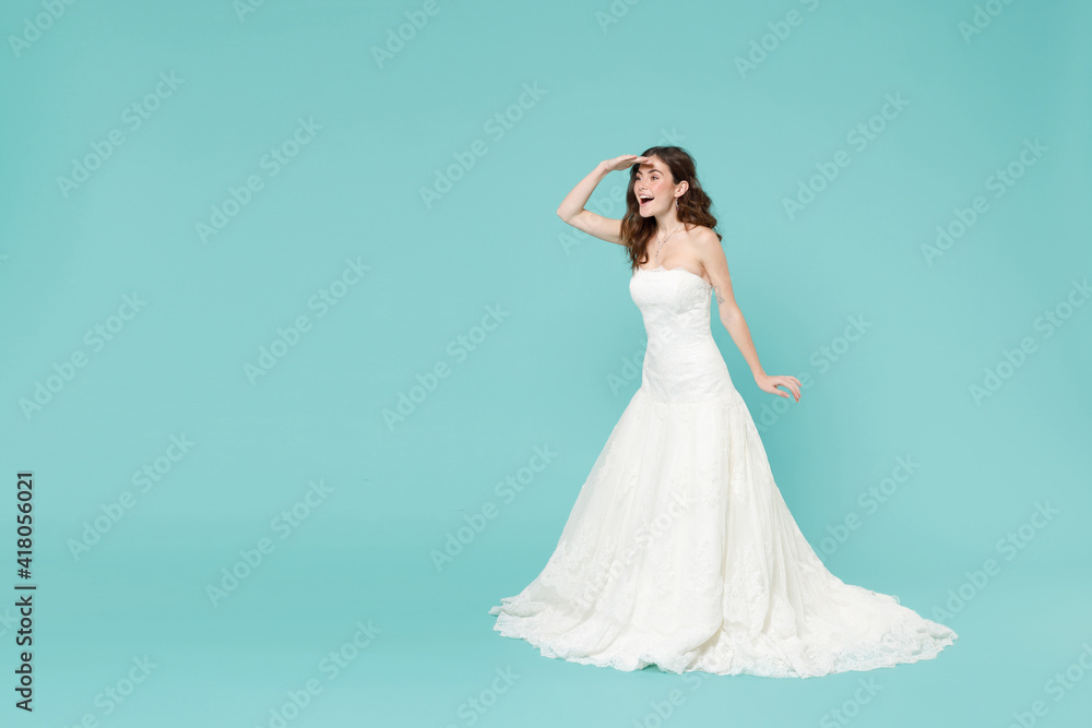 Full length of excited bride woman in white wedding dress holding hand at forehead looking far away distance isolated on blue turquoise background studio portrait. Ceremony celebration party concept. Full length of excited bride woman in white wedding dress holding hand at forehead looking far away distance isolated on blue turquoise background studio portrait. Ceremony celebration party concept.
