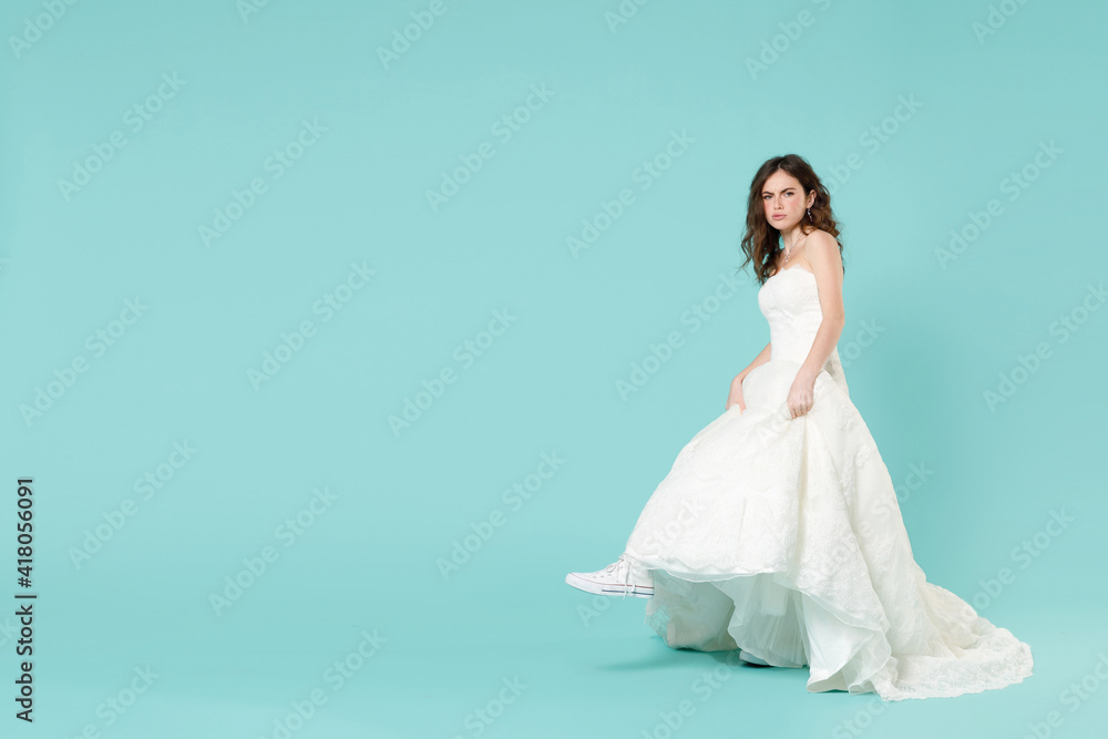 Full length side view of confused bride woman 20s in beautiful white wedding dress casual gumshoes looking camera isolated on blue turquoise background studio. Ceremony celebration party concept. Full length side view of confused bride woman 20s in beautiful white wedding dress casual gumshoes looking camera isolated on blue turquoise background studio. Ceremony celebration party concept.