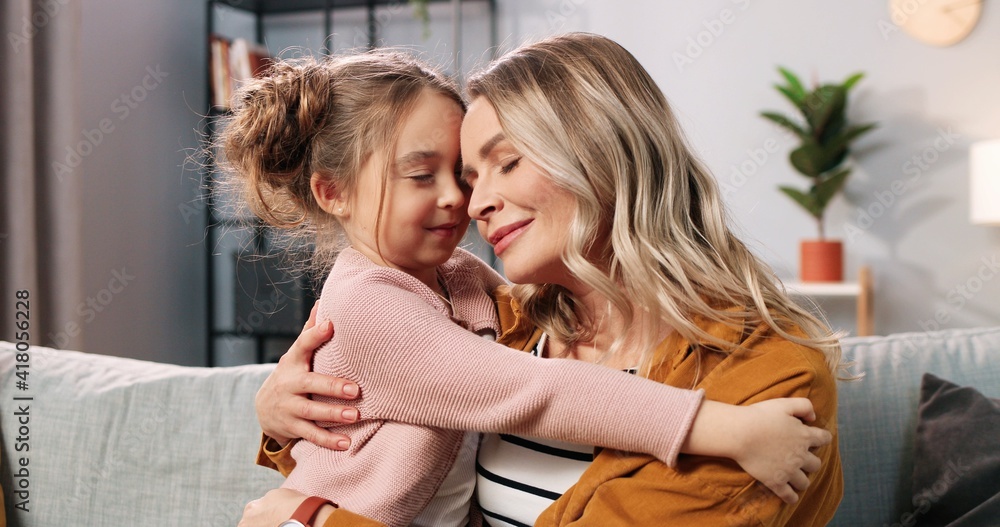 Close up of cute nice loving Caucasian little girl child hugging mother in living room at home. Caring mom embracing small daughter of sofa in apartment, love and care, family time concept Close up of cute nice loving Caucasian little girl child hugging mother in living room at home. Caring mom embracing small daughter of sofa in apartment, love and care, family time concept