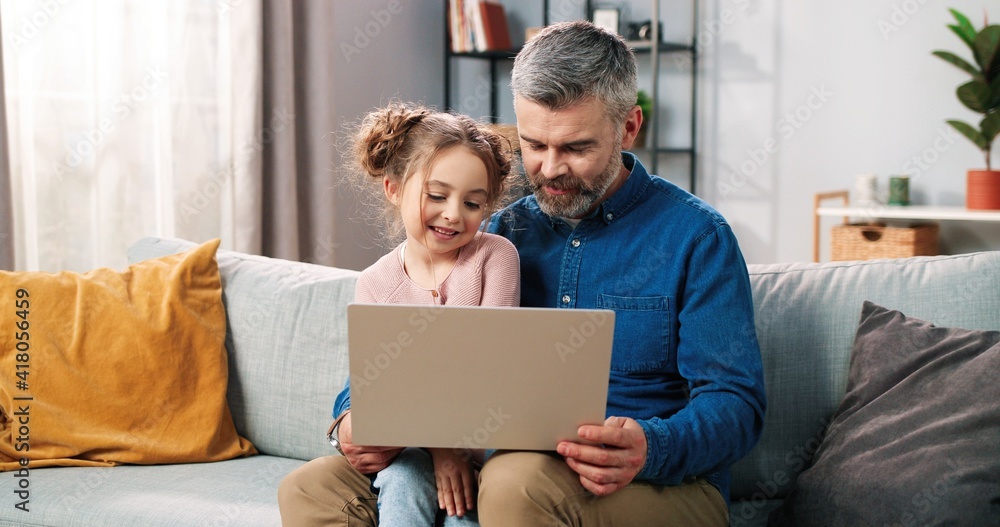 Portrait of happy nice Caucasian little daughter and caring father spending time together at home using laptop online. Dad and cute small kid girl tapping on computer browsing on internet