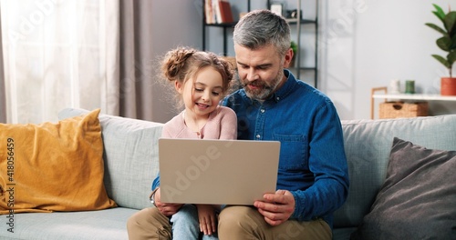 Portrait of happy nice Caucasian little daughter and caring father spending time together at home using laptop online. Dad and cute small kid girl tapping on computer browsing on internet