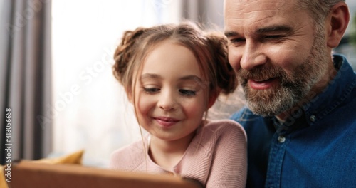 Close up of joyful caring Caucasian bearded dad with small daughter in room together videochatting using tablet. Father teaching small girl to use gadget device, family time, watching cartoons at home