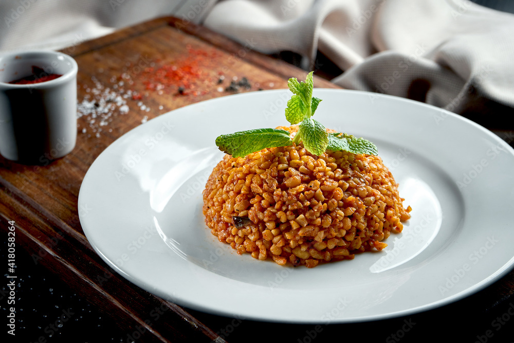 Appetizing and healthy side dish - bulgur in red sauce with Turkish spices in a white plate on a dark background