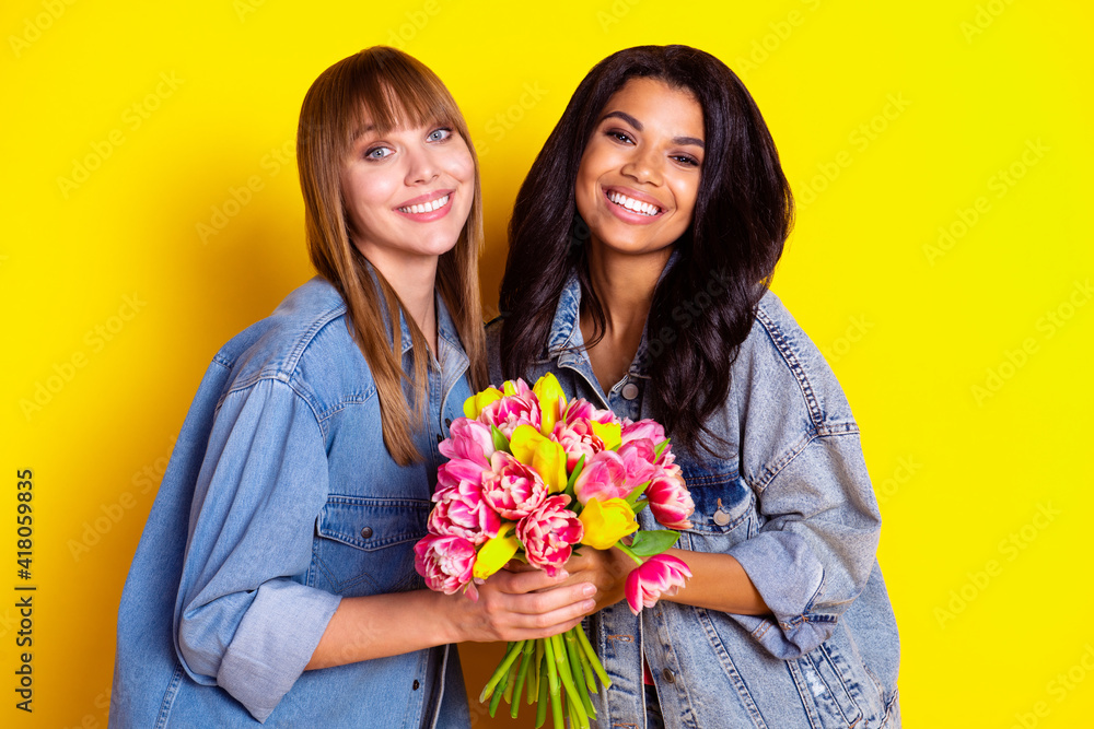 Portrait of two charming cheerful girls arms hold flowers toothy smile isolated on yellow color background Portrait of two charming cheerful girls arms hold flowers toothy smile isolated on yellow color background