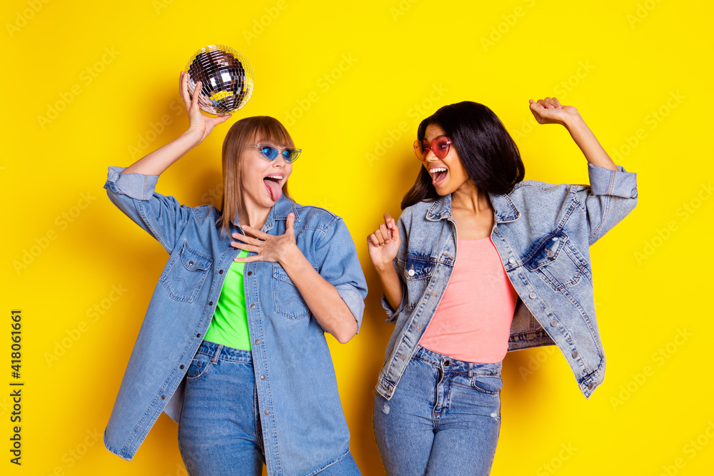 Portrait of two carefree girls arm hold disco ball open mouth tongue out have fun isolated on yellow color background