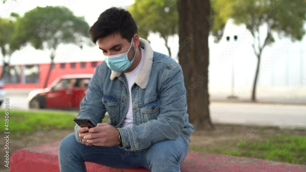Chico joven apuesto sentado en un banco con mascarilla por covid 19 usando el smartphone Chico joven apuesto sentado en un banco con mascarilla por covid 19 usando el smartphone
