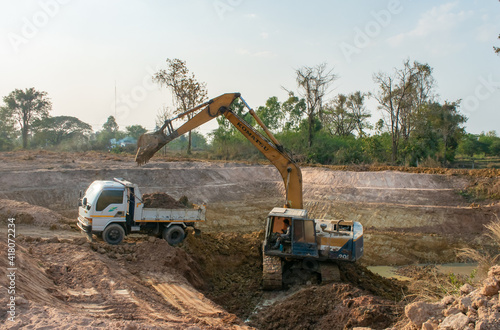 NAKHONRATCHASIMA-THAILAND-FEBUARY 27, 2021: Yellow excavator on the construction site loads the soil into the body of white dump truck, work on excavation to make a pond for agriculture.