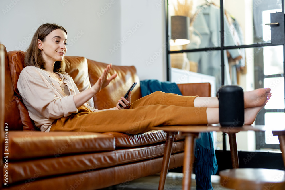 Obraz premium Happy pretty woman speaking with black audio assistant column on the coffee table while sitting on brown leather sofa in stylish living room. Smart home concept.