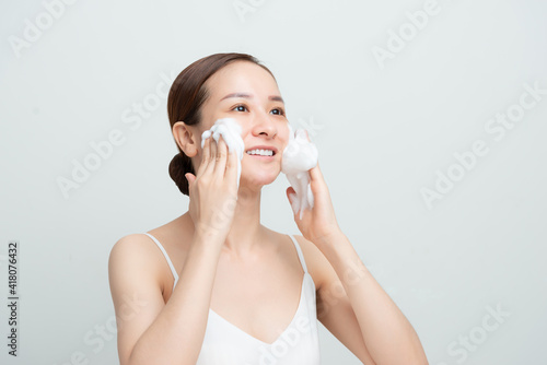 Satisfied beautiful woman makes massage of face, has white foam, cares of skin, smiles positively, models over white studio wall