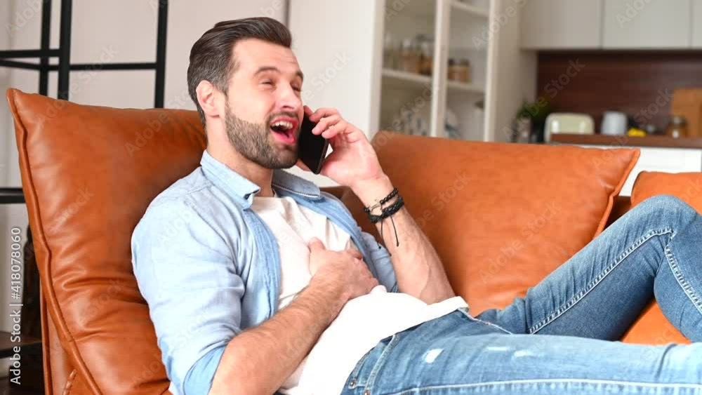 Attractive bearded hipster talks by the smartphone lying on the comfortable couch, smiling young man has phone conversation at home Attractive bearded hipster talks by the smartphone lying on the comfortable couch, smiling young man has phone conversation at home