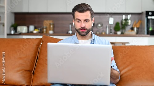 Front view of smiling young bearded hipster guy sitting on the couch, waving at laptop with high five, video-calling with family coworkers online, studying remotely, male freelnacer talking on webcam photo