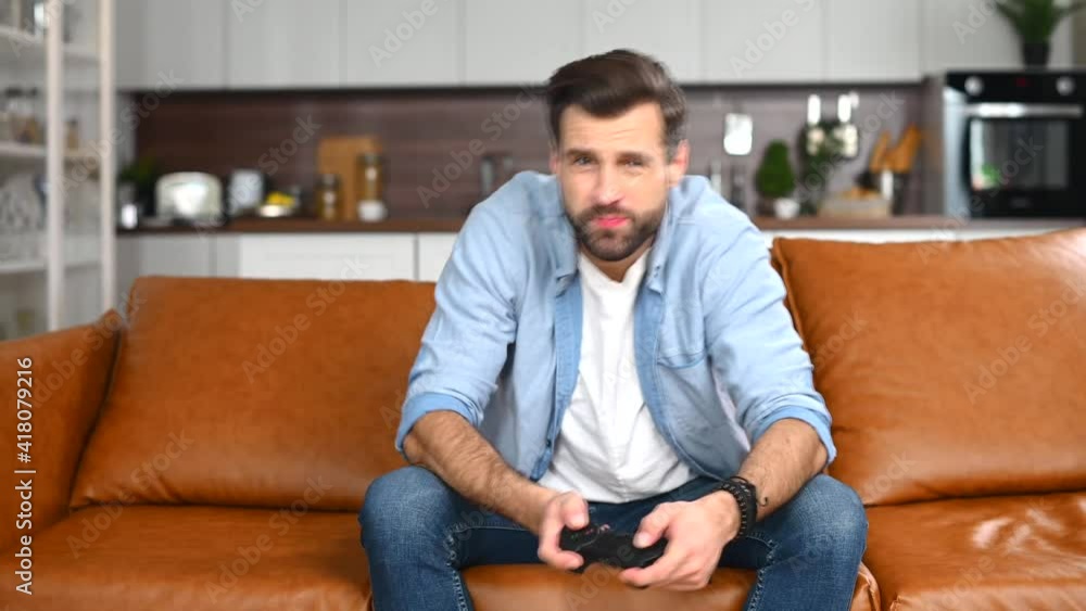 Front view portrait of disappointed young bearded hipster guy, gamer, or addict sitting on the couch in a living room, holding joystick, feeling stressed and worried because of the video game failure Front view portrait of disappointed young bearded hipster guy, gamer, or addict sitting on the couch in a living room, holding joystick, feeling stressed and worried because of the video game failure