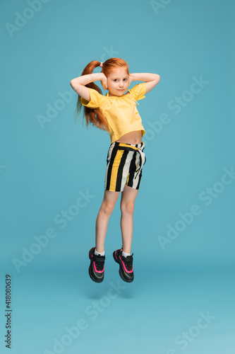 Fototapeta Naklejka Na Ścianę i Meble -  Jumping high. Happy, smiley little caucasian girl isolated on blue studio background with copyspace for ad. Looks happy, cheerful. Childhood, education, human emotions, facial expression concept.