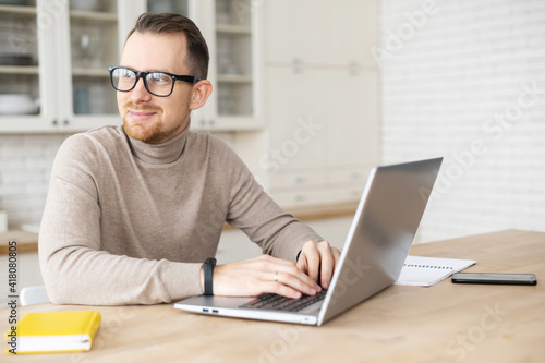 Handsome young hipster freelancer man with beard in glasses sitting at the table and looking away, taking a break from working on laptop, dreaming about vacation or salary, enjoying working from home