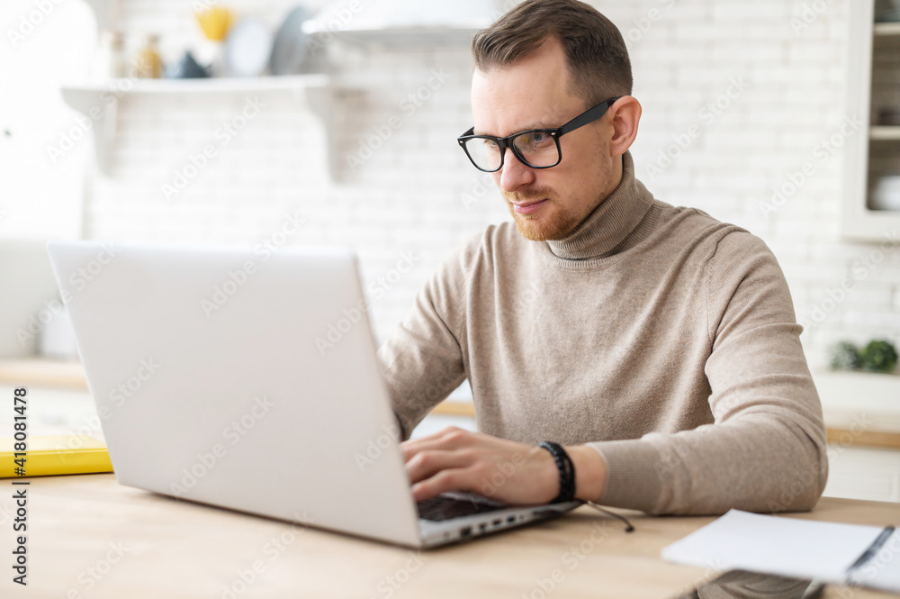 Serious focused determined young bearded businessman or freelancer in glasses and casual clothes sitting at kitchen table, typing on laptop, working on project, researching, writing email to client Serious focused determined young bearded businessman or freelancer in glasses and casual clothes sitting at kitchen table, typing on laptop, working on project, researching, writing email to client