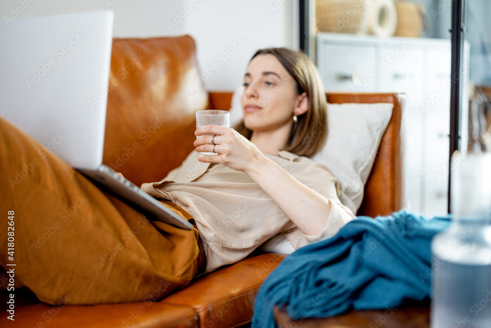Pretty woman hold a glass of water while working on laptop and lying on sofa. Lifestyle and healthy concept. Dehydration. Rate of drinking water per day Pretty woman hold a glass of water while working on laptop and lying on sofa. Lifestyle and healthy concept. Dehydration. Rate of drinking water per day