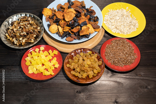 Dried fruits, oatmeal, flax seed, raisins, candied pineapple and walnuts lie on a dark background on multi-colored saucers.