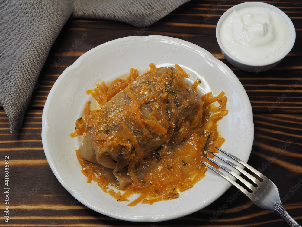 Vegetable roll with cabbage and carrots for vegetarian food in a plate, sour cream, fork on a wooden table, flat layout. Portion of a traditional dish golubtsy, golabki in sauce for a diet lunch