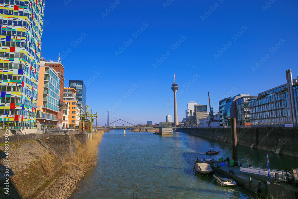 Obraz premium Düsseldorf, Germany - March 1. 2021: View over water canal beyond modern buildings on rhine tower against blue sky