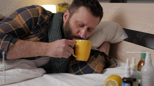 A sick man lying in bed home drinks hot herbal tea from yellow mug. Season of colds. bearded man of Caucasian nationality.