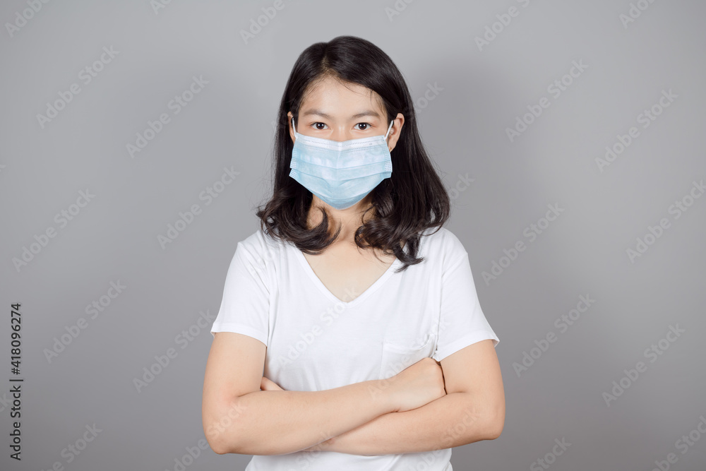 Portrait of Asian teenager in medical face mask to protect Covid-19 (Coronavirus) wears white t-shirt standing crossed arms isolated on gray background , health and medicine concept Portrait of Asian teenager in medical face mask to protect Covid-19 (Coronavirus) wears white t-shirt standing crossed arms isolated on gray background , health and medicine concept