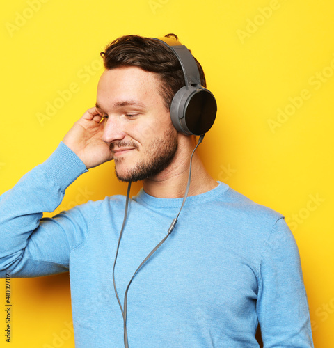 Young stylish man adjusting his headphones and smiling while standing against yellow background
