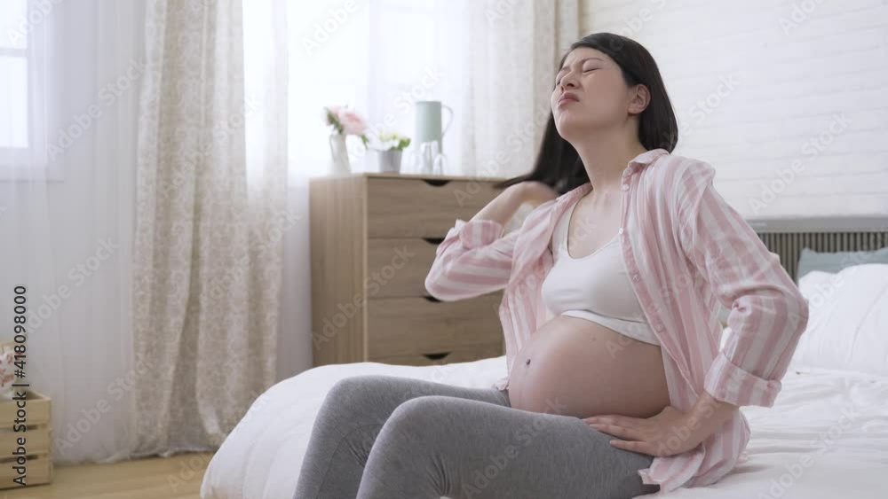 unhappy asian female suffering heatstroke during pregnancy is fanning herself and loosening her hair while sitting with an arm akimbo in the bedroom. unhappy asian female suffering heatstroke during pregnancy is fanning herself and loosening her hair while sitting with an arm akimbo in the bedroom.
