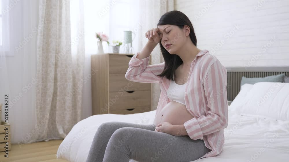 portrait asian expectant mom is feeling dizzy and rubbing her brows with discomfort on bed in the bedroom at home. portrait asian expectant mom is feeling dizzy and rubbing her brows with discomfort on bed in the bedroom at home.