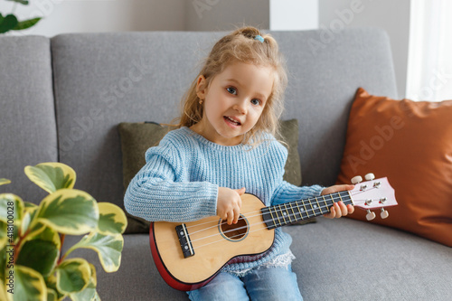 Portrait of cute little girl playing ukulele and singing it at home. 3 years old kid learning guitar. Concept of early childhood education, music hobby, talent, happy preschool leisure and parenting