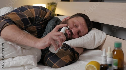 A sick person lies in bed at home, he has sore throat, laryngitis, tonsillitis. A man sprays an aniseptic spray down his throat. Season of colds, fever.
