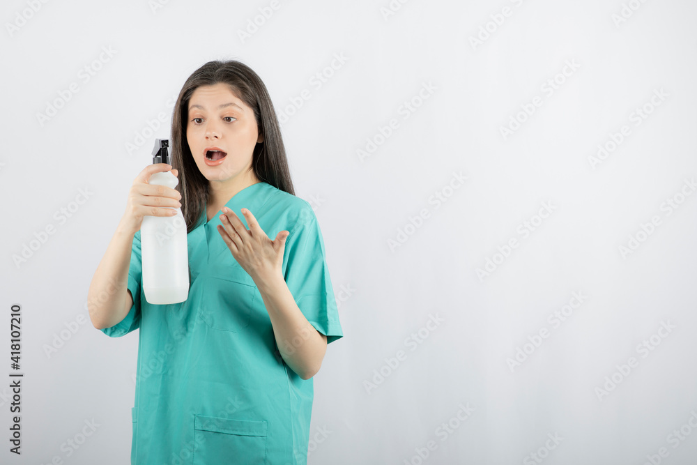 Shocked young nurse looking at disinfectant bottle and making hand gesture Shocked young nurse looking at disinfectant bottle and making hand gesture