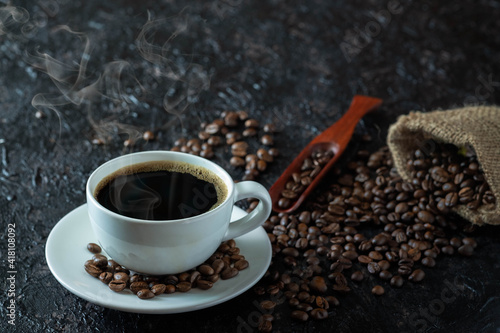 Hot black coffee with smoke for morning beverage menu in white ceramic cup with coffee beans roasted in burlap sack bag on dark grunge rustic table background. Flat lay with copy space.