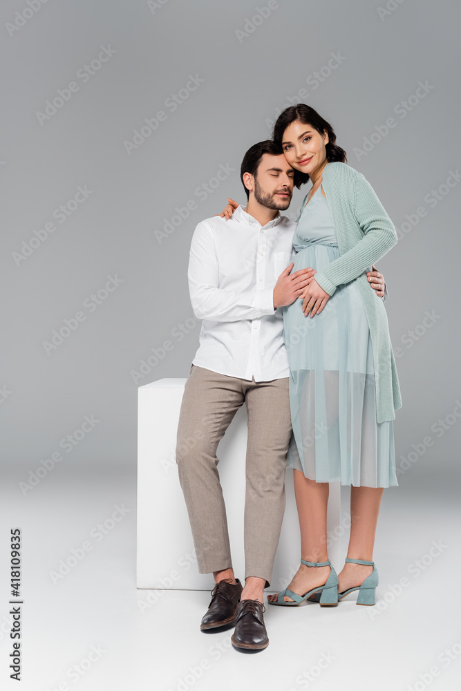 Young man embracing stylish pregnant wife near white cube on grey background Young man embracing stylish pregnant wife near white cube on grey background