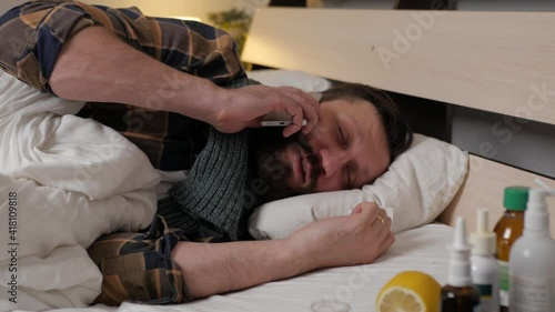 A sick man is lying in bed home, talking phone and wiping his nose with handkerchief. The man is upset and has difficulty communicating, his throat hurts. Season of colds, fever, infection, flu.