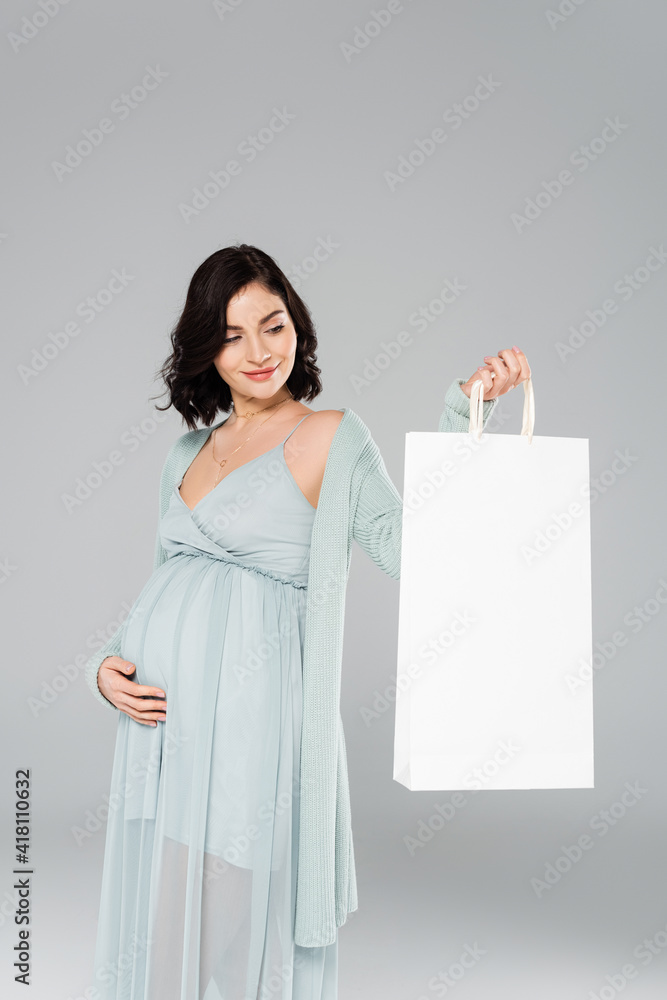 Smiling pregnant woman looking at shopping bag isolated on grey Smiling pregnant woman looking at shopping bag isolated on grey