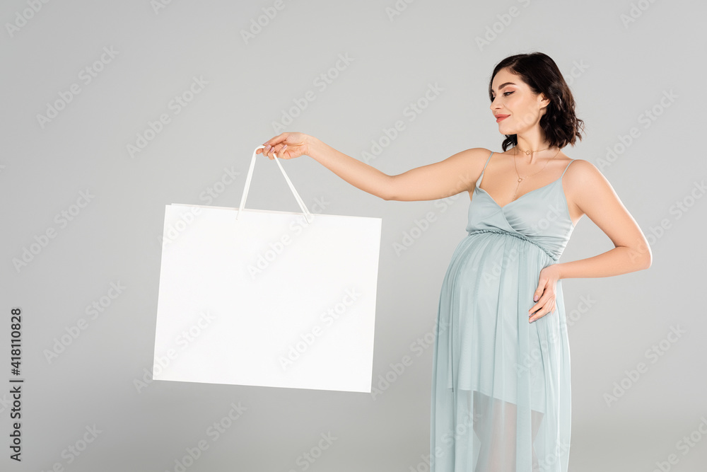 Pregnant woman in dress looking at white shopping bag isolated on grey Pregnant woman in dress looking at white shopping bag isolated on grey