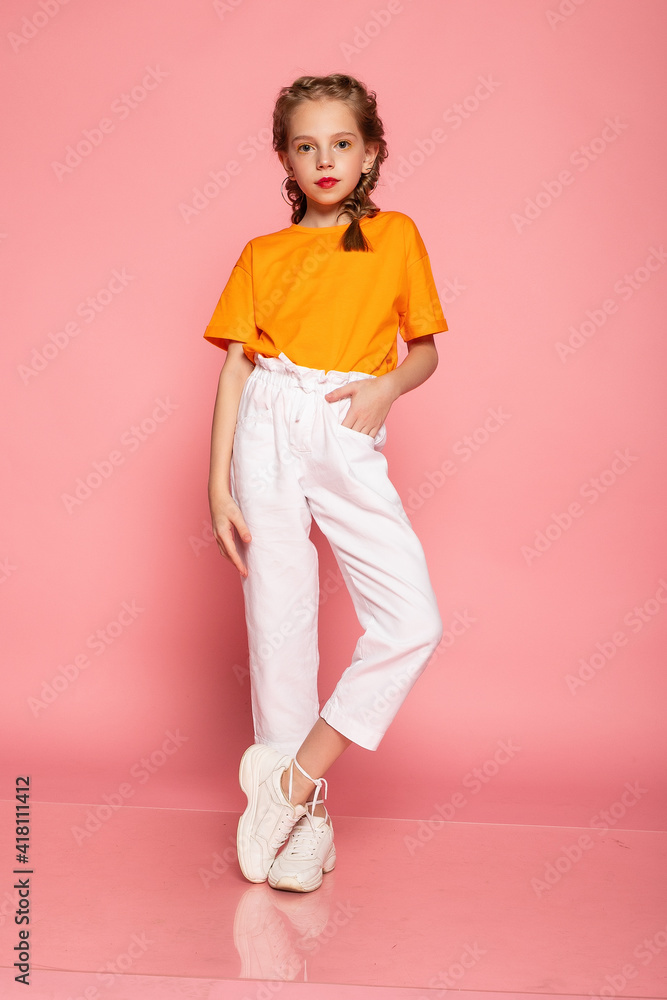 full length little girl on pink studio background. wearing an orange T-shirt and white pants and white sneakers. Stylish kids, happy childhood, emotions. copy space