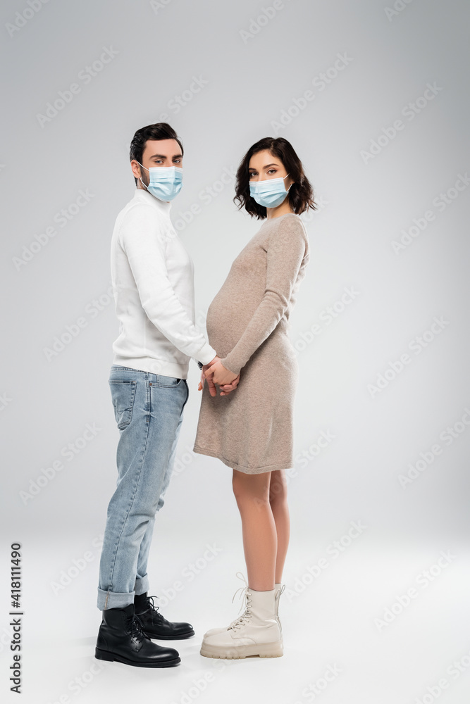 Pregnant woman in medical mask holding hand of husband on grey background Pregnant woman in medical mask holding hand of husband on grey background