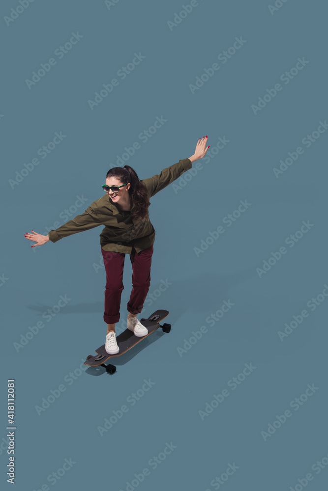 Skateboarding. High angle view of young woman on blue studio background. Girl in motion. Human emotions and facial expressions concept. Full length portait, copyspace for ad. Fashion, retro style.