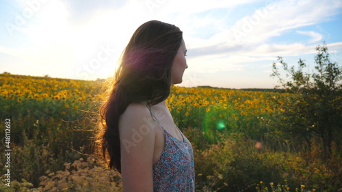 Profile of pretty girl walking outdoor with blooming sunflower field on background. Young woman going through countryside enjoying freedom and beautiful summer environment at sunset. Slow motion