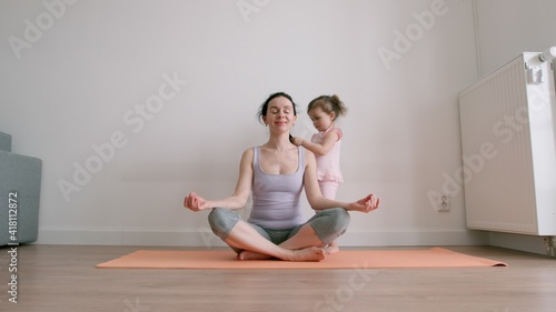 Mother sits in lotus position on yoga mat and little baby girl plays with her mother's hair at home