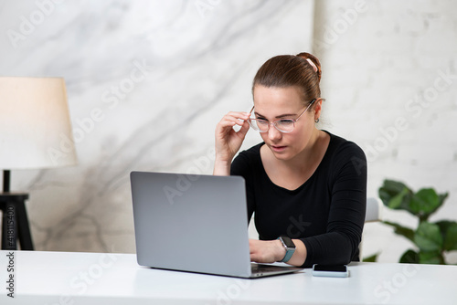 Young concentrated focused girl, serious beautiful woman in glasses is typing, working on her laptop computer at table at home with eyesight problems, hardly trying to read online content at screen. 