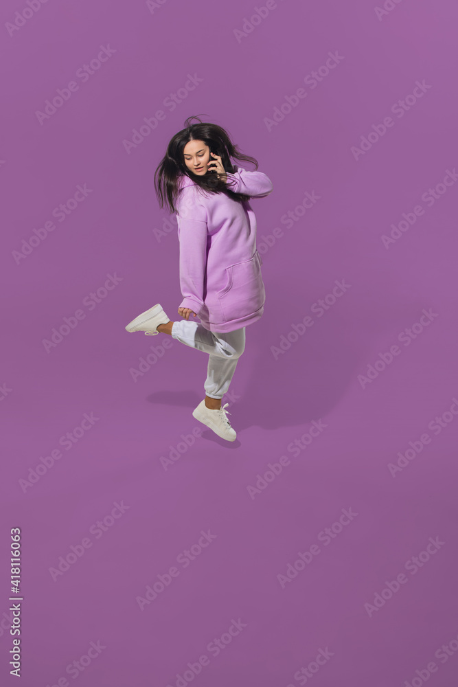Talking phone. High angle view of young woman on purple studio background. Girl in motion. Human emotions and facial expressions concept. Full length portait, copyspace for ad. Fashion, retro style.