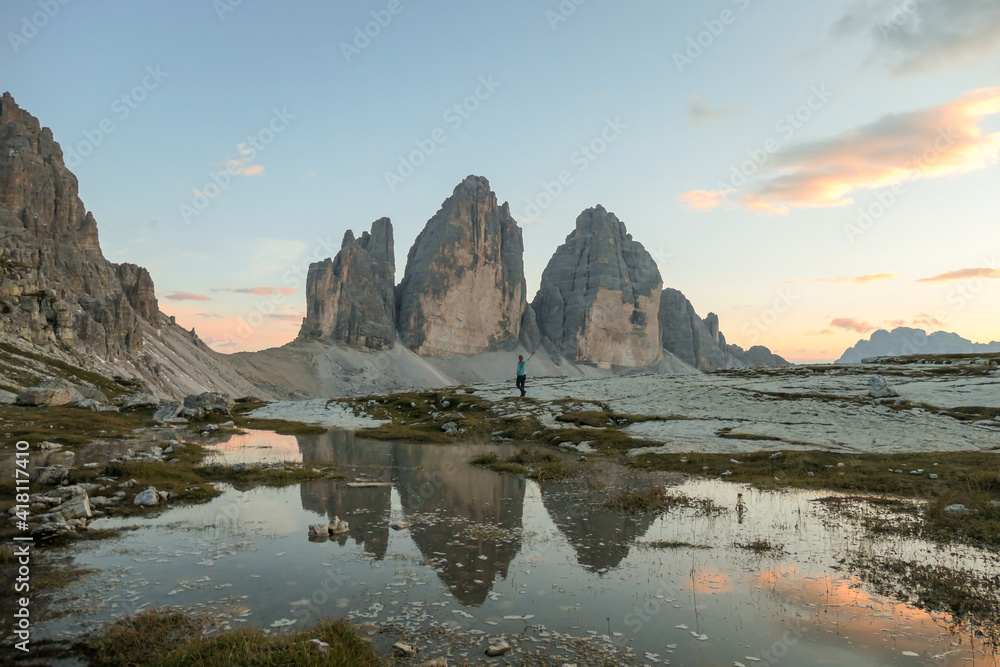 A woman enjoying the sunset over the Tre Cime di Lavaredo (Drei Zinnen) mountains in Italian Dolomites. The peaks reflect in a paddle. The mountains are surrounded with orange and pink clouds. Freedom