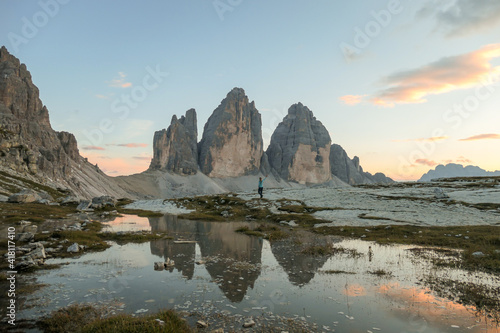 A woman enjoying the sunset over the Tre Cime di Lavaredo (Drei Zinnen) mountains in Italian Dolomites. The peaks reflect in a paddle. The mountains are surrounded with orange and pink clouds. Freedom
